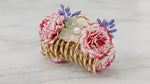 Gold metal coil claw clip with two full pink-red carnation blooms, white lace tulle rosette with pearl centre and purple lavender berry clusters on marble surface